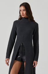 A woman is wearing a long, dark gray, ribbed knit sweater with a high neckline. The sweater has a slit from the waist down, revealing black leather shorts underneath. The sweater's design includes vertical ribbing and a semi-open front from the waist down.